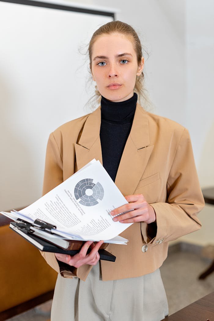Caucasian woman in a business suit holding documents, standing confidently in an office setting.